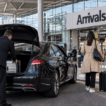 A black minicab is parked in Gatwick Airport’s North Terminal drop-off area while a man loads a suitcase into the boot and a woman walks towards the Arrivals entrance with luggage