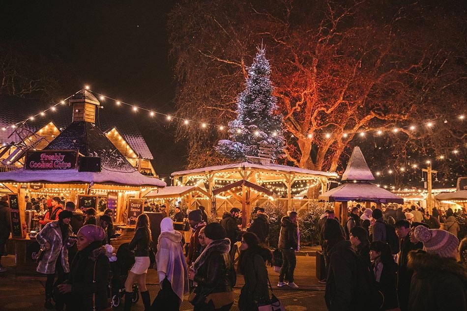 Hyde Park Winter Wonderland: Visitors enjoying Hyde Park Winter Wonderland in London, surrounded by festive food stalls, Christmas trees and twinkling lights.