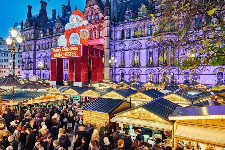 Manchester Christmas Markets: Wooden Christmas market stalls decorated with festive lights and a giant illuminated Santa in the background